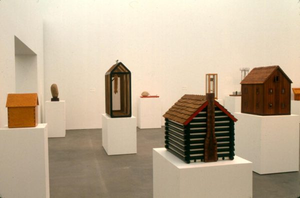 Small 3-d houses of varying sizes and styles on white pedestals in a white gallery space