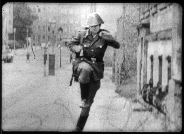 Archival image of a person dressed in a vintage military uniform and helmet running down a sidewalk