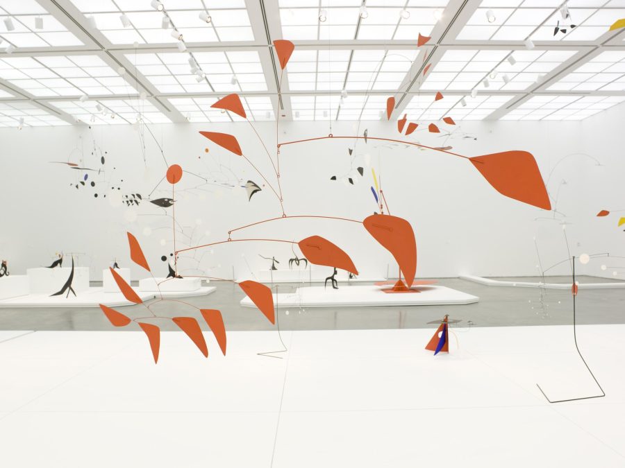 Interior view of a brightly lit gallery featuring an extensive display of Alexander Calder's sculptures, characterized by their abstract shapes and vibrant colors. At the forefront is an orange mobile suspended in air.