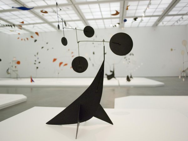 A black sculpture balances a mobile of circles in a white gallery spaces filled with other mobiles