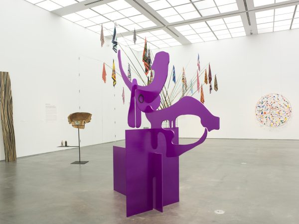 An art gallery installation featuring a large purple abstract sculpture in the foreground, with various colorful hanging artworks and other sculptural pieces in the background.