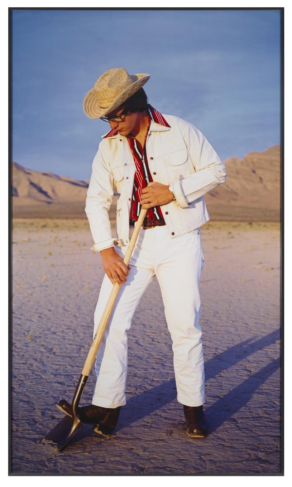 A man in a white jean jacket and pants wearing a cowboy hat holds a shovel to the ground in a desert setting