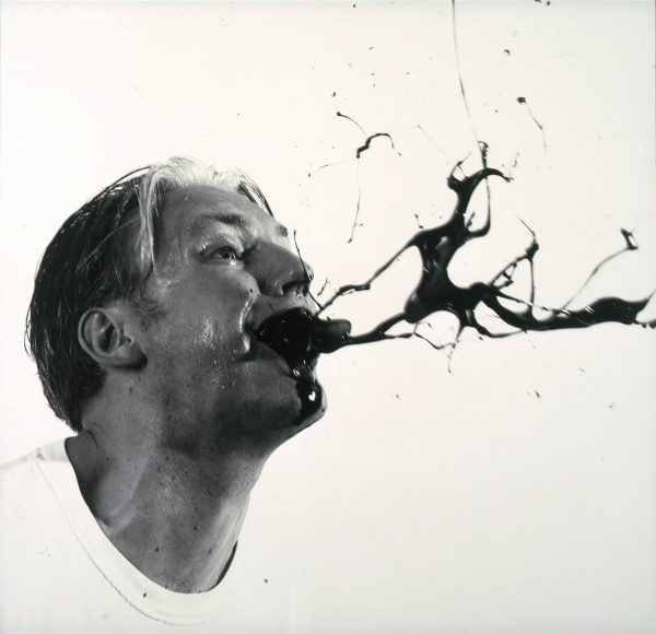 Black and white photo of a man spitting out black liquid. Their white t-shirt almost blends into the white background making it look like their head is floating.