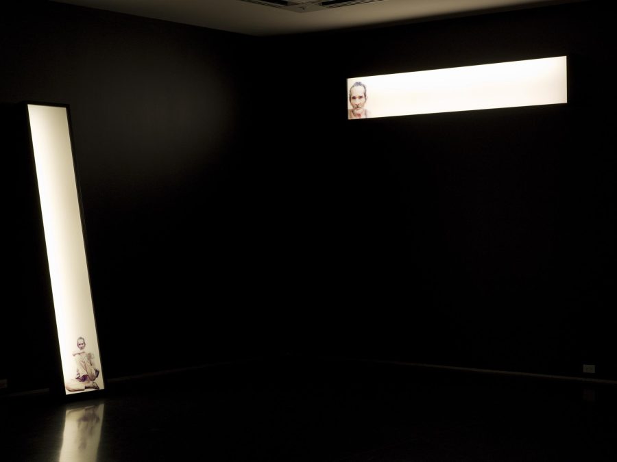 Two narrow white glowing rectangles depicting a man illuminate a pitch-black room. The horizontal box on the right hangs high on the wall; the other leans against the wall to the left.
