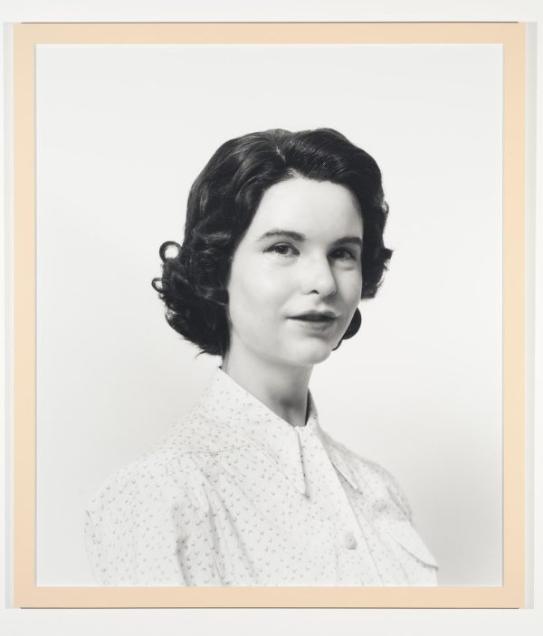 A black-and-white photograph portrait shows a pale-skinned person with short dark hair wearing a white blouse in front of a white background.