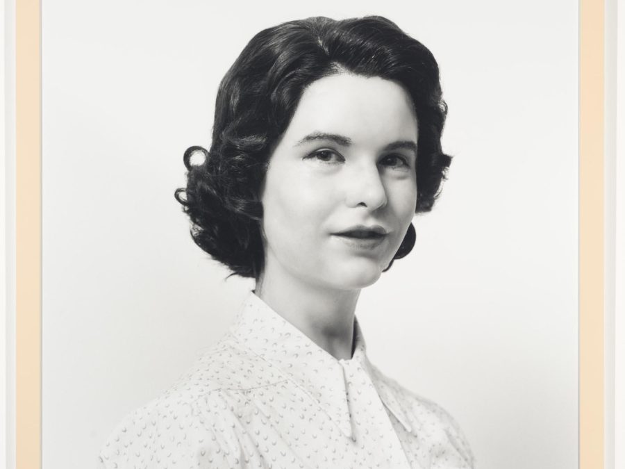 A black-and-white photograph portrait shows a pale-skinned person with short dark hair wearing a white blouse in front of a white background.