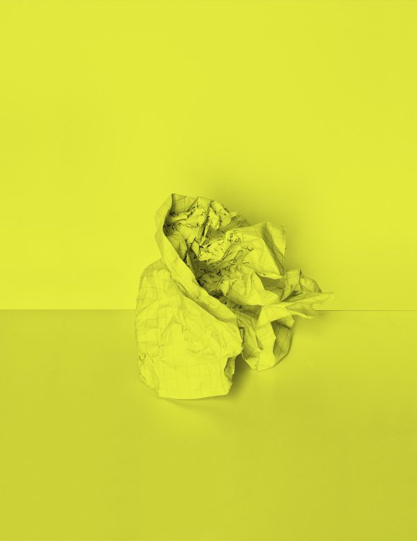 A lime green wall serves as a backdrop for a lime green ball of crumbled graph paper. The paper has words scribbled on it, which are indistinguishable due to how heavily it has been crumbled.