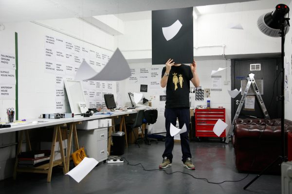 A person holds a black posterboard in front of their face in an office space with white printer paper floating around them