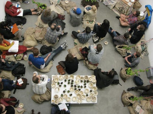 Overhead view of a large group of people sitting on a concrete floor facing away from a table filled with small bowls