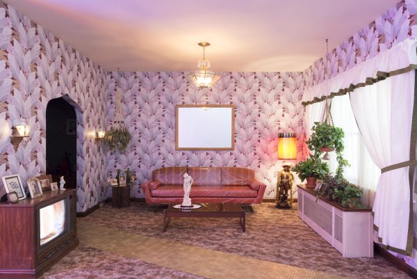A 1970s style living room with floral trellis style wallpaper and furnished with a couch, coffee table, box tv, side tables, and lamps. A doorway to the left leads to a pitch-black room.