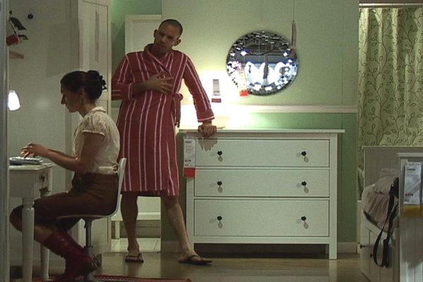 A person in a red and white striped robe leans on a white dresser while looking at a person sitting in a white chair at a table