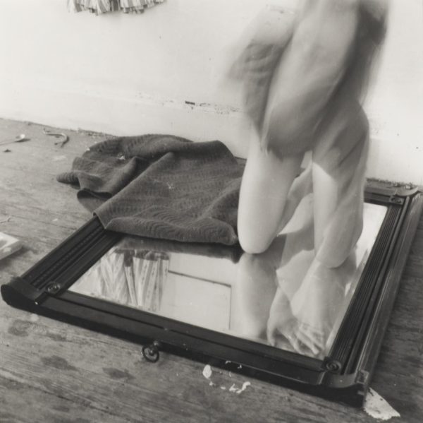 A black-and-white photograph shows a pale-skinned person kneeling on a mirror placed on the floor with a towel covering part of the mirror. The top part of the person's body is in motion and is blurry.