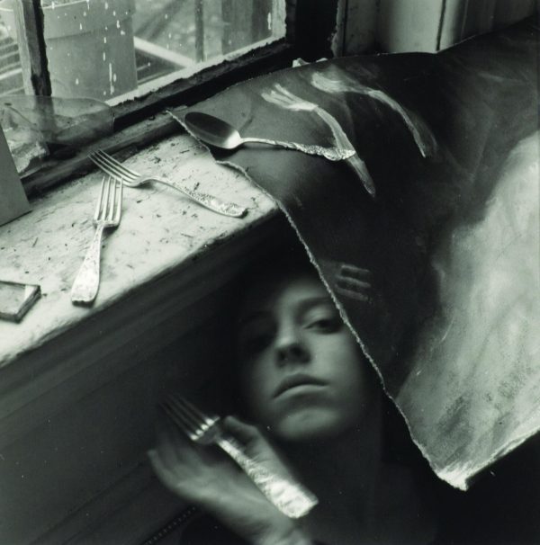 An obstructed face peers at the viewer from under a canvas sheet with forks strewn about.