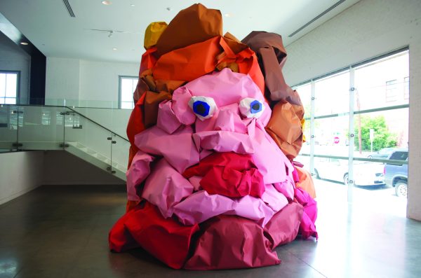 A giant blob with eyes and red lips made out of pink, red, and brown paper in a modern space with a glass staircase behind it