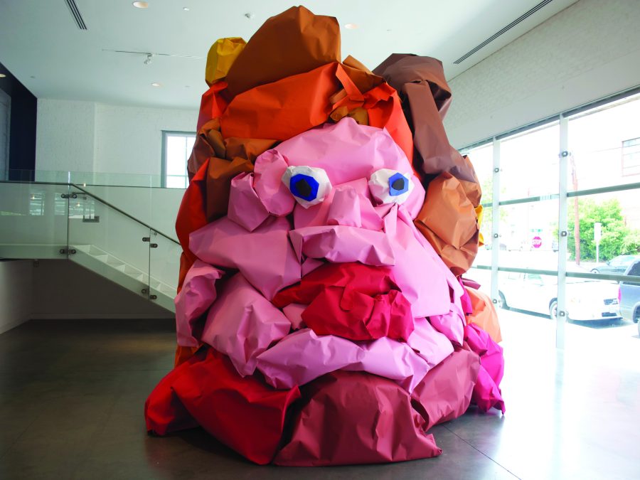 A giant blob with eyes and red lips made out of pink, red, and brown paper in a modern space with a glass staircase behind it
