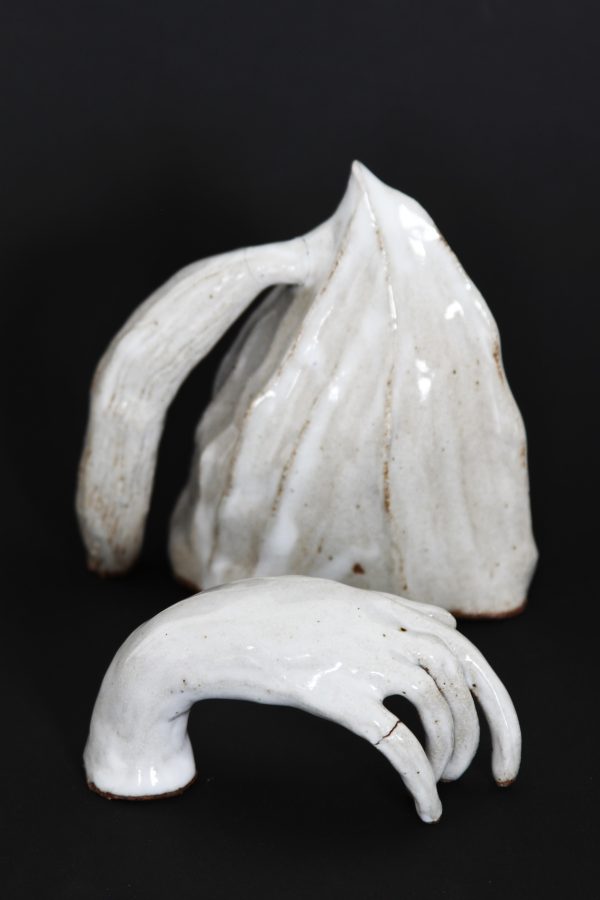 A white pyramid-shaped ceramic sculpture has a long white handle attached. A white ceramic hand with a visible crack in the pinky finger sits in front of the other sculpture.