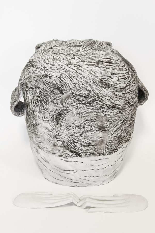 A white ceramic vessel bears black, scratch-like markings. A pair of white ceramic hands lays in front of the vessel.