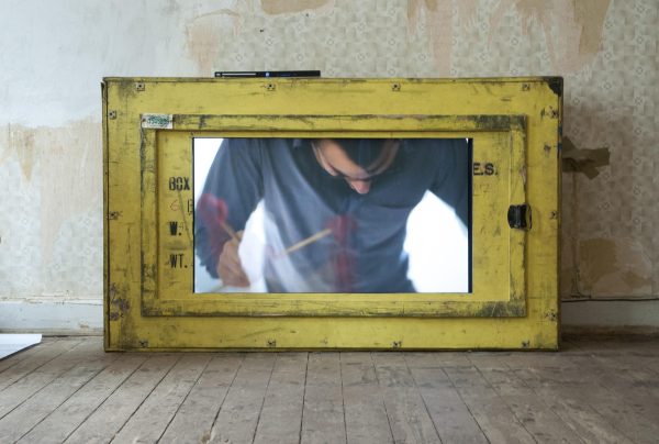 A person hunched over is viewed from a small yellow window at the base of a wall