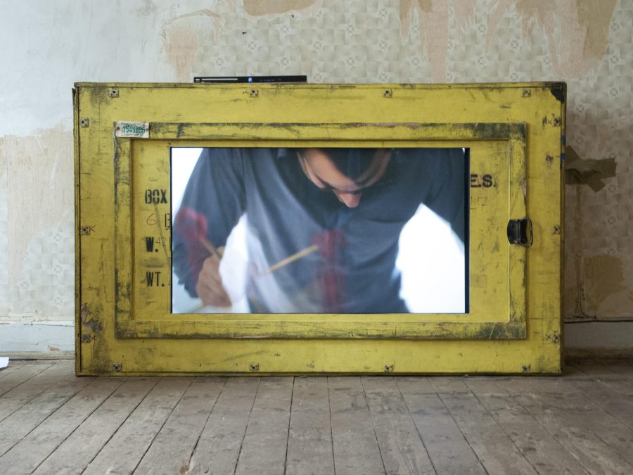 A person hunched over is viewed from a small yellow window at the base of a wall
