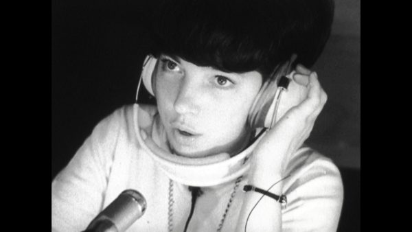 Black and white image of a female-presenting person holding headphones up to their left ear while speaking into a small microphone