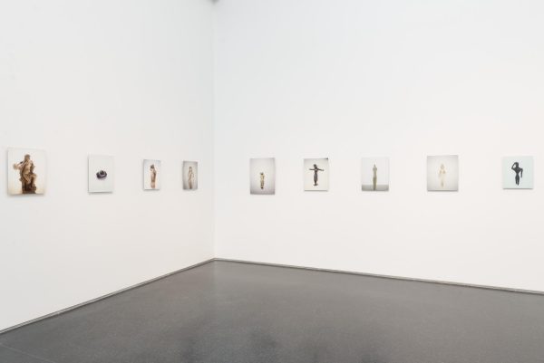 Nine photographs are hung on a white wall. Each photograph has a different sculpture from various civilizations throughout history.