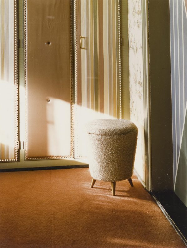 A small upholstered stool in the corner of a 1960s style room is illuminated by a band of sunlight