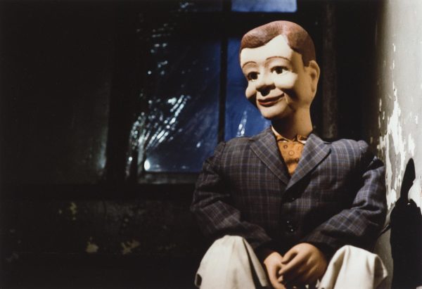 A male ventriloquist puppet sits in a darkened staircase wearing a plaid blazer and khaki pants.