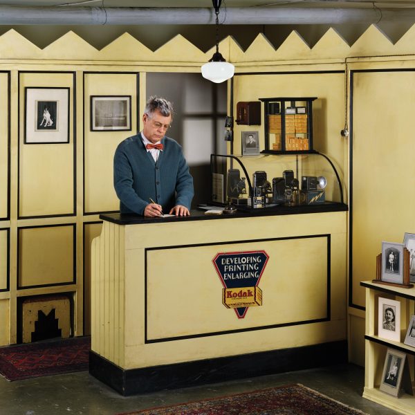 A man in a cardigan and bowtie stands behind the counter of a small booth. A Kodak advertisement reads 'developing printing enlarging'