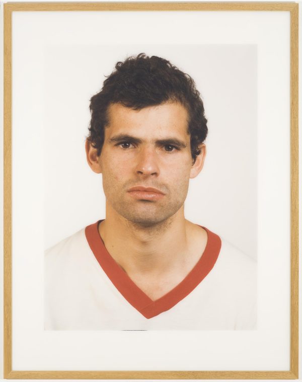 Portrait of an unsmiling male-presenting person with short curly dark hair in a v-neck white t-shirt with a red neckband