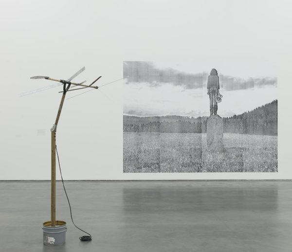 A large white gallery space with grey concrete floors features an antennae in a paint can in the middle of the room, near the foreground of the image, and a greyscale image of a person standing on a plinth in the middle of a field on the wall