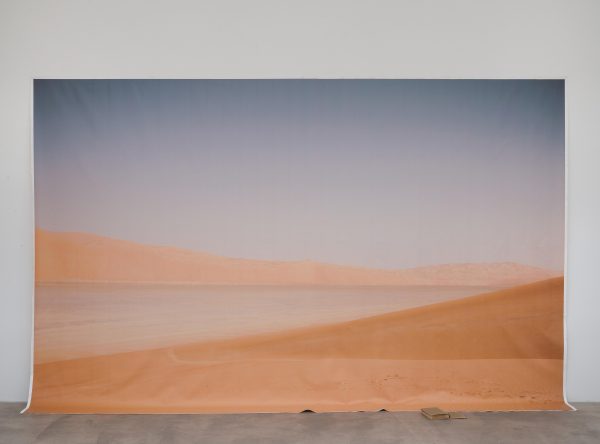 Picture of a desert landscape mostly hung on the white wall with a small portion cascading onto the floor