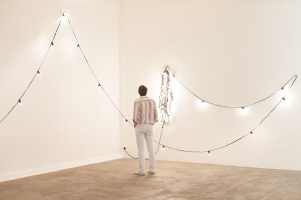 A person is standing in the corner of a room with their back to you. A string of white lights is hung loosely on the walls.