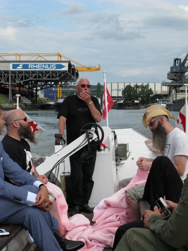 A boat tour with the captain standing at the helm holding a handheld microphone to his mouth. Next two him are two men sitting across from each other. One is knitting a large pink object.