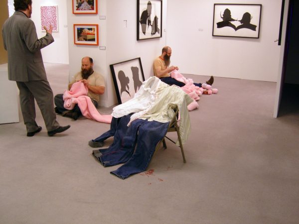 In a gallery space filled with art hanging on the walls two male-presenting people sit on the ground on two sides of a corner knitting something pink.