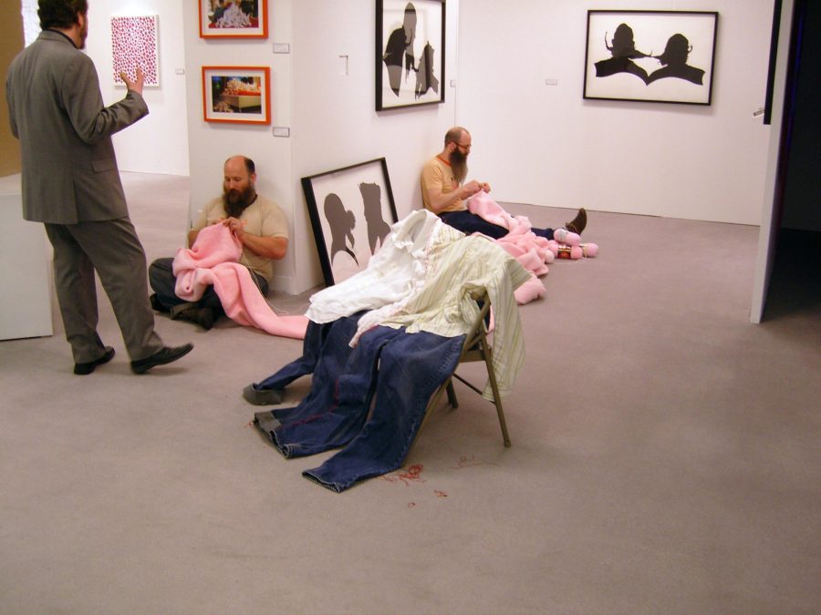 In a gallery space filled with art hanging on the walls two male-presenting people sit on the ground on two sides of a corner knitting something pink.