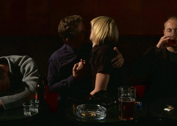 Two people kissing in a dark bar setting with two people to either side of them. One is drinking, the other is passed out on the table.