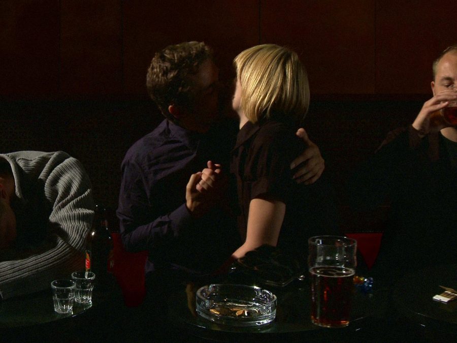 Two people kissing in a dark bar setting with two people to either side of them. One is drinking, the other is passed out on the table.