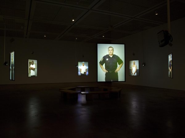 Screens hanging in the middle of a dark room display images of various people posing against a gray screen. There is a circle of benches in the middle of the room.