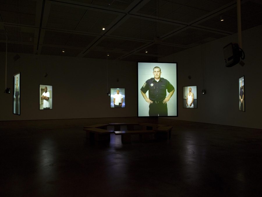 Screens hanging in the middle of a dark room display images of various people posing against a gray screen. There is a circle of benches in the middle of the room.