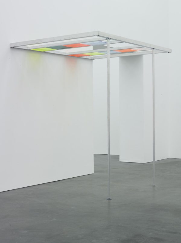 A pergola-like structure held up by two thin metal poles in a white-walled room. The roof of the structure is divided into four rows, two of which have various colored light filters