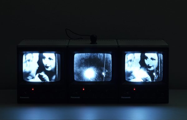 Three tv screens illuminate a dark room. The two on the left and right display the same image of a woman; the middle displays a white orb on the bottom.