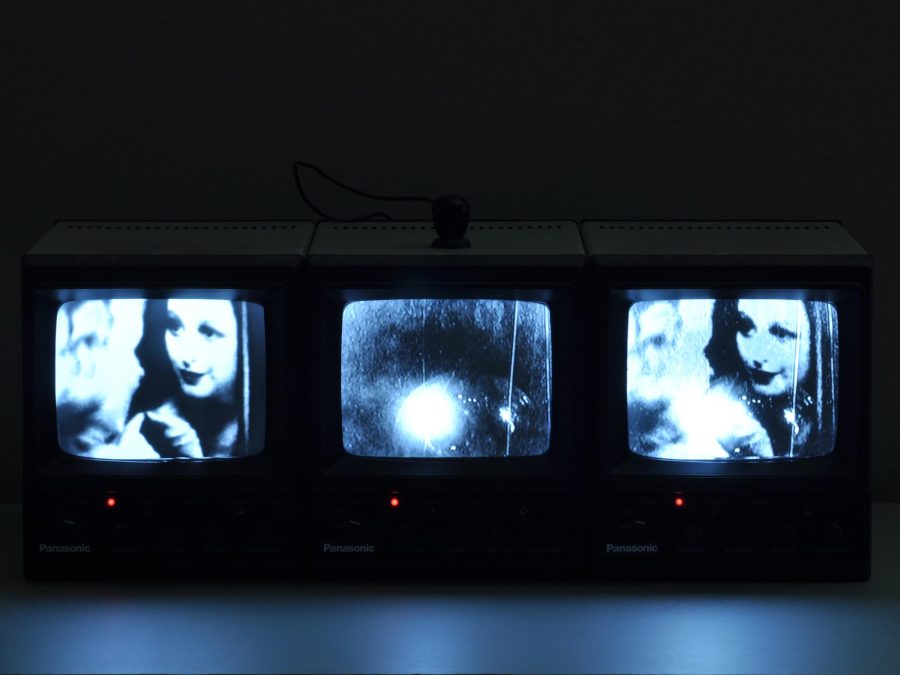 Three tv screens illuminate a dark room. The two on the left and right display the same image of a woman; the middle displays a white orb on the bottom.