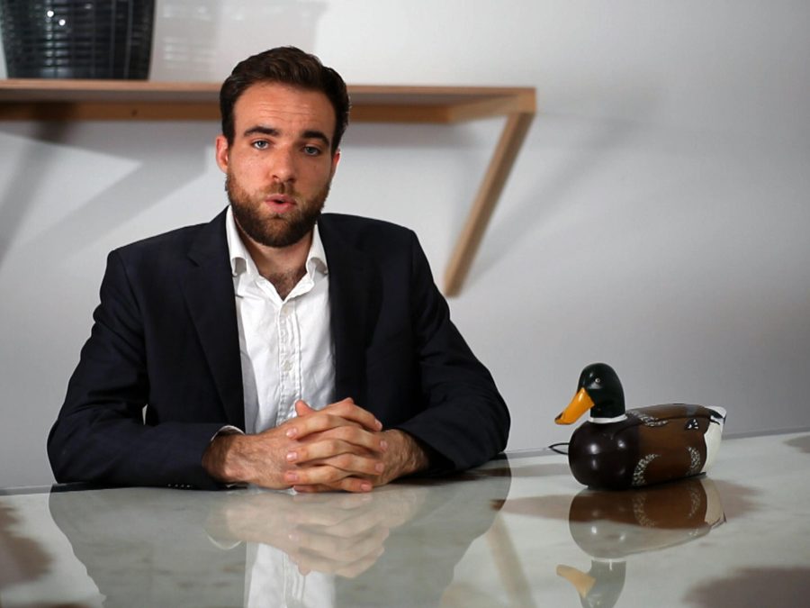 A man in a suit jacket sits at a table with hands folded in front of him and a duck phone to his left. His lips are pursed, as if in mid sentence.