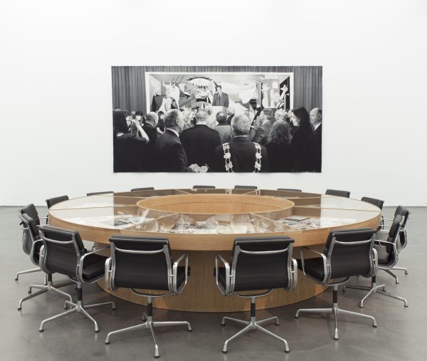 Office chairs surround a circular wooden table with glass on top. An artwork of a group of people looking at an artwork hangs on a white wall behind it.