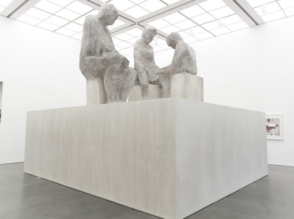 A giant grey pedestal features a larger-than-life sculpture of three grey figures seated together in a brightly lit white gallery space