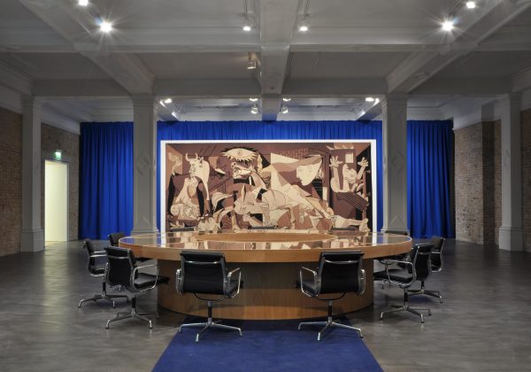 A round table surrounded by office chairs in front of Pablo Picasso's Guernica against blue curtains