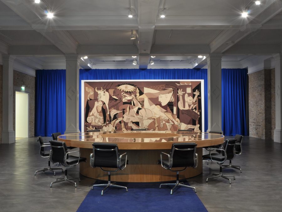 A round table surrounded by office chairs in front of Pablo Picasso's Guernica against blue curtains