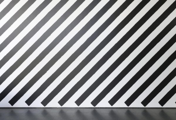 Black stripes painted vertically on a white wall from upper right to bottom left, ending just before the floor