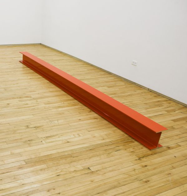 An orange building beam lays in a room with a wooden floor and white walls.