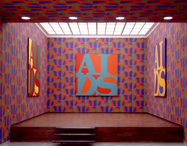 An elevated room covered in red, blue, and green repeating wallpaper on three walls that matches the art which features the acronym AIDS in the style of the Robert Indiana LOVE artwork on the three walls.